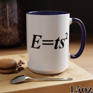 May include: White ceramic mug with a navy blue interior and handle. The mug features the equation E=ts² in black lettering. The mug is labeled "15oz". A spoon, coffee beans, and a macaron are on a wooden surface.