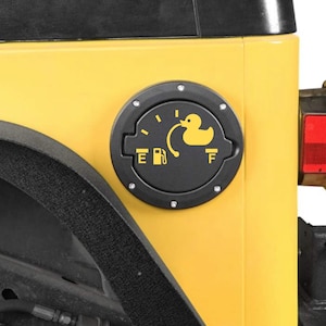 May include: A black fuel door cover with a yellow rubber ducky graphic on a yellow vehicle. The graphic includes a fuel gauge and the letters "E" and "F". The cover is round and has silver bolts around the edge.