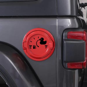 May include: A circular red fuel door cover for a vehicle featuring a black graphic decal. The decal displays a fuel gauge design with an E and F, a gas pump icon, and a black rubber duck silhouette positioned near the full mark of the gauge.