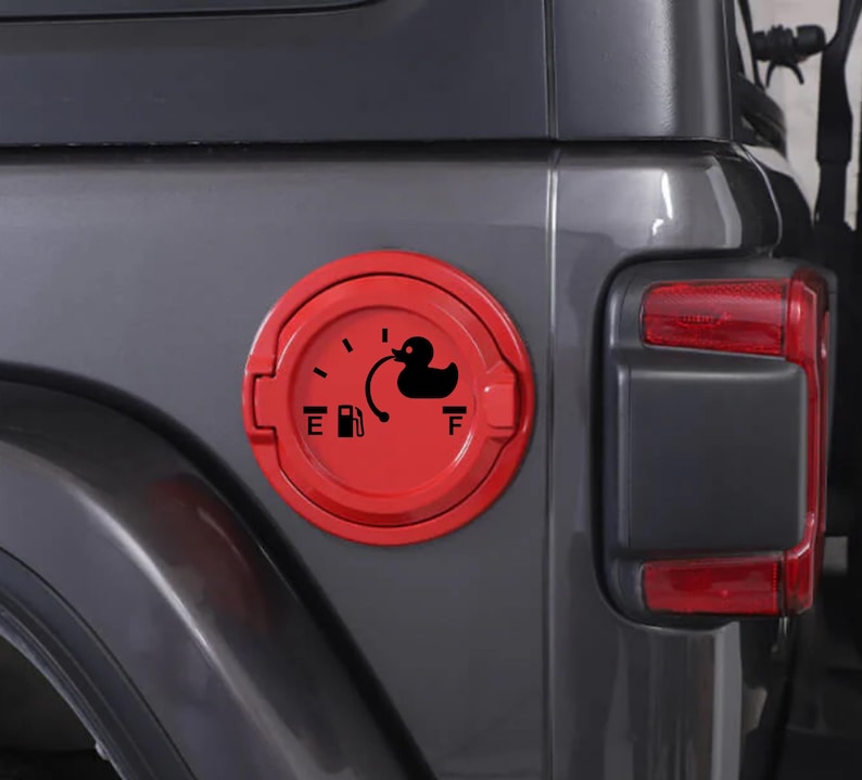 May include: A circular red fuel door cover for a vehicle featuring a black graphic decal. The decal displays a fuel gauge design with an E and F, a gas pump icon, and a black rubber duck silhouette positioned near the full mark of the gauge.