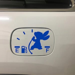 May include: A blue decal featuring a cartoon character holding a gas pump nozzle, designed to look like a fuel gauge. The gauge shows the fuel level between "E" and "F". The decal is on a white car.