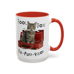 May include: A white ceramic mug with a red interior and handle. The mug features a tabby cat holding a wrench in its mouth, sitting in a red toolbox. The text on the mug reads "Tool Box" and "Su-Purr-visor".
