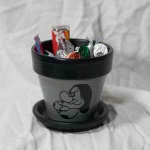 May include: A gray ceramic flower pot with a black rim and a matching saucer. The pot features a black graphic of a cartoon character and is filled with wrapped candies.