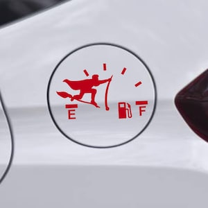 May include: Red vinyl decal depicting a figure on a broomstick, with a cape, above a fuel gauge. The gauge shows the fuel level near empty. The decal is on a white car.