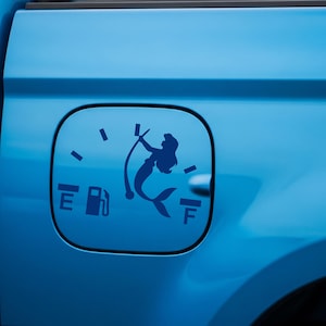May include: Blue car fuel door with a blue mermaid graphic. The graphic includes a fuel gauge, a mermaid holding a bow, and the letters "E" and "F".