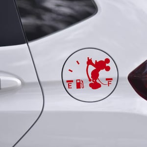 May include: Red Mickey Mouse fuel gauge sticker on a white car. The sticker is a playful design, featuring Mickey Mouse holding the fuel gauge needle. The gauge shows the fuel level at empty. The car's door handle and tail light are also visible.