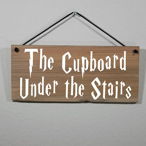 May include: A wooden sign with the text "The Cupboard Under the Stairs" in white lettering. The sign is hanging from a black string.