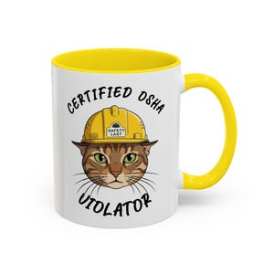 May include: White ceramic mug with a yellow interior and handle. The mug features a cartoon cat wearing a yellow hard hat, with the words "CERTIFIED OSHA VIOLATOR" printed on the front.