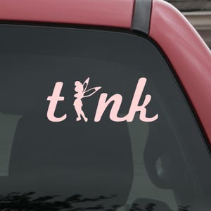 May include: Pink decal with the word "tink" in a whimsical font, featuring a silhouette of a fairy replacing the "i". The decal is displayed on a car window, with the car's red paint visible.
