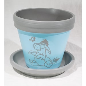 May include: A light blue ceramic flower pot with a gray rim and matching saucer. The pot features a gray illustration of a donkey and a butterfly. A decorative item for plants.