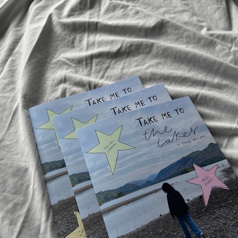 K&ouml;nnte beinhalten: Drei Exemplare eines Zines mit dem Titel "Take Me To The Lakes" von Ahsia Patel. Das Cover zeigt einen blauen Himmel, ein Gew&auml;sser und eine Person, die an einem felsigen Ufer steht. Das Zine hat einen gelben Stern mit den Worten "Digitalkamera" und einen rosa Stern mit den Worten "Speiseplan".