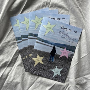 K&ouml;nnte beinhalten: Ein Stapel Zines mit dem Titel "Take Me To The Lakes" mit einer handgezeichneten Illustration einer Person, die auf einem Strand mit einem See im Hintergrund steht. Das Zine hat einen rosa Stern mit dem Text "Real Plans" und einen gelben Stern mit dem Text "What We Wanted".