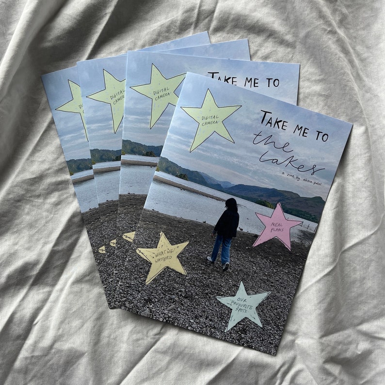 K&ouml;nnte beinhalten: Ein Stapel Zines mit dem Titel "Take Me To The Lakes" mit einer handgezeichneten Illustration einer Person, die auf einem Strand mit einem See im Hintergrund steht. Das Zine hat einen rosa Stern mit dem Text "Real Plans" und einen gelben Stern mit dem Text "What We Wanted".
