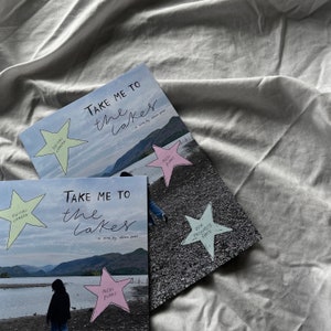 K&ouml;nnte beinhalten: Zwei Exemplare eines Zines mit dem Titel "Take Me To The Lakes" von Ahsia Patel. Das Zine zeigt Fotos eines Sees und von Bergen. Die Cover sind mit handgezeichneten Sternen und Text verziert.