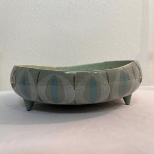 May include: A light blue ceramic bowl with a white and blue abstract design. The bowl has three legs and is slightly oval shaped.