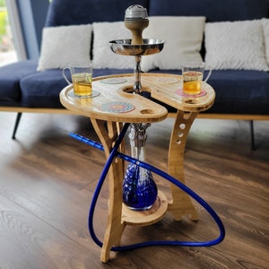 Könnte beinhalten: Ein Shisha-Tisch aus Bambus mit blauem Glasfuß und silberfarbenen Metallakzenten. Der Tisch hat zwei Holzoberflächen mit kreisförmigen Aussparungen für Getränke und dekorative Untersetzer. Zwei Gläser mit bernsteinfarbener Flüssigkeit stehen auf dem Tisch, mit einem blauen Schlauch.