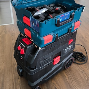 Könnte beinhalten: Ein schwarz-roter Staubsauger mit einer blau-roten Werkzeugbox oben drauf. Der Staubsauger hat ein Stromkabel und Räder. Die Werkzeugbox hat einen Griff und ein Etikett mit der Aufschrift "Bosch".