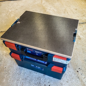 Peut inclure: Une boîte à outils bleue et rouge avec un dessus noir. Le dessus est en bois et a une surface texturée. La boîte porte l'inscription "Bosch Professional 18V GSR 18V-140 FC" sur le côté.