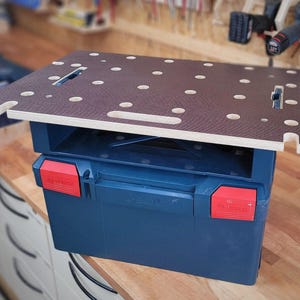 Peut inclure: Une boîte à outils bleue avec un loquet rouge et une surface de travail en bois brun avec des trous sur le dessus. La surface de travail mesure environ 46 cm sur 61 cm.