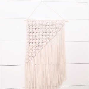 Peut inclure: Une suspension murale en macramé blanche avec un motif géométrique et des franges. La suspension est faite de cordon de coton et est fixée à une baguette en bois.