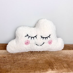 Op de afbeelding: Een wit, pluche wolkvormig knuffeldier met geborduurde ogen, een glimlachende mond en roze bloemaccenten.
