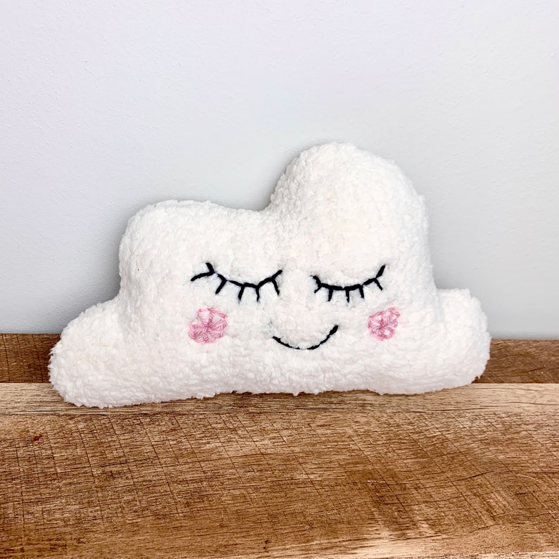 Op de afbeelding: Een wit, pluche wolkvormig knuffeldier met geborduurde ogen, een glimlachende mond en roze bloemaccenten.