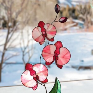 Puede incluir: Escultura de orquídea de vidrieras con pétalos rosas y rojos, hoja verde y base negra con acento morado. La escultura se encuentra sobre un fondo borroso de paisaje invernal.