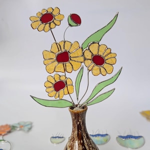 Puede incluir: Un arreglo floral decorativo de vidrio en un jarrón de cerámica marrón y azul. Las flores son amarillas con centros rojos y hojas verdes. Flores de vidrio adicionales de varios colores están dispersas.