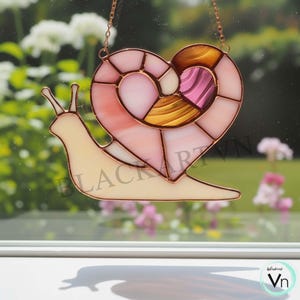 Peut inclure: Un capteur de soleil en vitrail en forme d'escargot avec une coquille en forme de cœur. La coquille présente des morceaux de verre roses, blancs et dorés. Le corps de l'escargot est de couleur jaune pâle et la pièce est suspendue à une chaîne délicate.