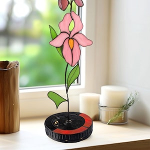 Puede incluir: Una escultura de flores de vidriera con una orquídea rosa con hojas y capullos verdes. La flor está montada sobre una base negra con un acento rojo. La escultura se exhibe en un alféizar de madera.