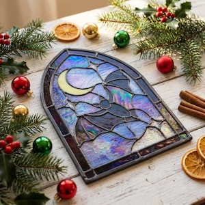 May include: An arched stained glass panel featuring a crescent moon and a bat silhouette. The glass pieces are iridescent, displaying shades of blue, purple, and white, held together by dark metal solder lines in a gothic window frame design.