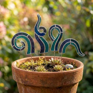 May include: Four stained glass garden stakes in shades of teal and dark blue, featuring swirling and arched shapes. These decorative pieces are inserted into a terracotta pot filled with soil and small succulent plants, creating a garden display.