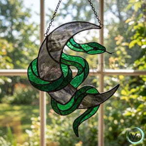 Op de afbeelding: Een glas-in-lood zonnevanger met een groene slang die om een halve maan van getextureerd grijs glas is gewikkeld. Het object is geconstrueerd met donkere metalen soldeerlijnen en hangt aan een dunne metalen ketting.
