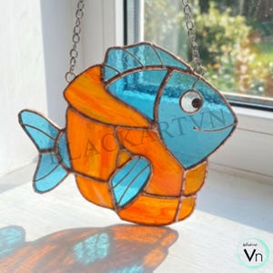 Könnte beinhalten: Buntes Glasfisch-Ornament mit orangefarbenen und blauen Segmenten, aufgehängt an einer silbernen Kette. Der Fisch hat ein lächelndes Gesicht und den Text "BLACKARTVN". Der Fisch wird vor einem Fenster präsentiert.