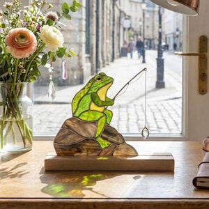 Peut inclure: Une figurine en vitrail représentant une grenouille verte assise sur un rocher brun, tenant une fine canne à pêche en métal avec un petit pendentif en cristal transparent suspendu au fil. L'objet est fixé sur un socle en bois clair.