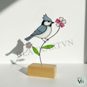 May include: A stained glass sculpture of a blue jay perched on a metal stem with a pink and white flower and green leaves. The bird is mounted on a small wooden block. The sculpture casts a shadow on the white background.