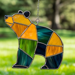 May include: A stained glass bear suncatcher with a silver metal frame. The bear is made of yellow, orange, green, and blue-green glass pieces. A silver chain is attached for hanging. The bear is a decorative item.