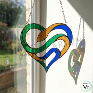 Puede incluir: Un atrapasoles de vidrio manchado en forma de corazón con un diseño de serpiente. El corazón presenta vidrio verde, azul y amarillo, suspendido por una cadena. El atrapasoles proyecta una sombra colorida en la pared. El diseño incluye el texto "blockart Vn".