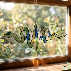 Può includere: Decorazione in vetro colorato con un design a forma di serpente e battito cardiaco, nei toni del verde, blu e trasparente. L'opera è sospesa davanti a una finestra, mostrando i dettagli e i colori. Un elemento decorativo.