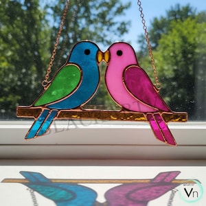 Könnte beinhalten: Glasmalerei mit zwei sich küssenden Vögeln. Ein Vogel ist blau und grün, der andere rosa und magenta. Sie sitzen auf einem braunen Ast, der an einer Kette hängt. Das Kunstwerk wird vor einem Fenster gezeigt.
