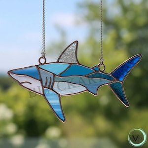 May include: A stained glass shark suncatcher in shades of blue, turquoise, and white, with a copper-coloured frame. The shark is suspended by a silver chain, ideal for a window or wall decoration. The design features geometric shapes.