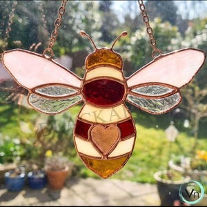 May include: A stained glass bee suncatcher, suspended by a copper chain. The bee design incorporates a heart and is crafted from pink, red, yellow, and clear glass. The word "BLACK" is partially visible.