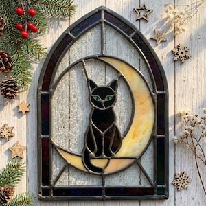 May include: A stained glass panel in an arched window frame featuring a black cat sitting on a pale yellow crescent moon. The cat has bright green eyes and is constructed with dark metal lead lines. The panel is surrounded by pine branches and stars.