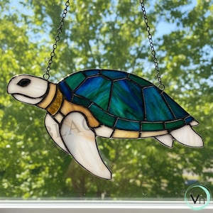 May include: Stained glass turtle suncatcher with a blue and green shell, white flippers, and a cream-colored head. The turtle is suspended by a black chain. The suncatcher is displayed in front of a blurred green background.