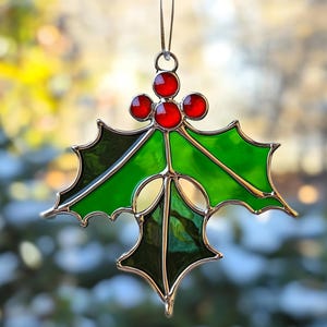 May include: A stained glass holly ornament with red berries and green leaves. The ornament is outlined in a metallic gold color and has a brown string for hanging. The ornament is a festive decoration.