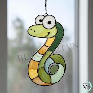 May include: A colourful stained glass snake suncatcher. The snake features a cheerful design with green, yellow, and orange segments, large white eyes, and a smiling face. It hangs from a chain, suitable for window or wall decor.