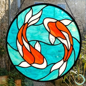 May include: A round stained glass panel featuring two koi fish in shades of orange, white, and black, set against a turquoise background. The fish are arranged in a circular pattern, creating a sense of movement and balance. The panel is suspended by a chain.