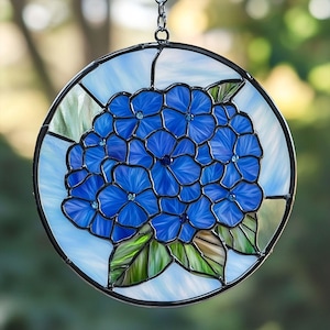 May include: Round stained glass window featuring a cluster of blue hydrangea flowers with green leaves. The flowers have small blue accents. The window is framed with a black border and a chain for hanging. The background is a light blue and white.