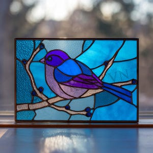 Peut inclure: Panneau en vitrail représentant un oiseau bleu perché sur une branche avec des baies. L'oiseau est rendu dans des tons de violet et de bleu, sur un fond de verre bleu texturé. Le panneau est encadré de métal foncé.
