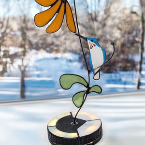 May include: A stained glass flower sculpture with an orange flower, green leaves, and a blue and white bird. The sculpture sits atop a round, black and white base with a light. The background is a snowy winter scene.